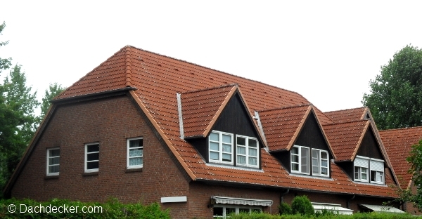 Mehrfamilienhaus mit rotem Ziegeldach in Deutschland. Das Dach weist mehrere gleichmäßig angeordnete Gauben mit dunkler Holzverkleidung und weißen Fenstern auf. Die Fassade besteht aus rotem Klinkermauerwerk, umgeben von dichter Begrünung.