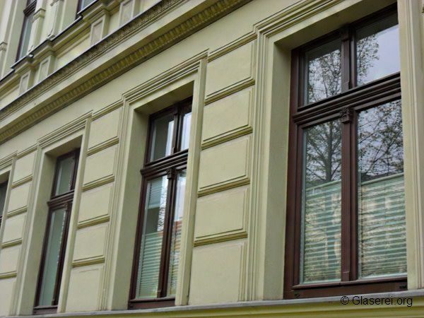 Fassade eines Gebäudes mit drei großen, dunkel umrahmten Fenstern. Die cremefarbene Wand hat dekorative Streifen und zeigt Reflexionen von Bäumen und das Blau des Himmels in den Fenstern. Veranstaltung urheberrechtlicher Angaben unten rechts.