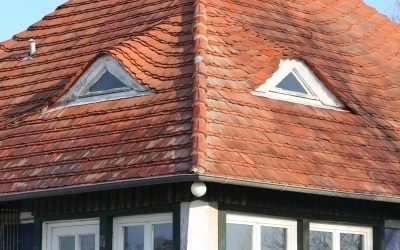 Haus mit stark geneigtem Walmdach aus roten Tonziegeln und zwei markanten Gaubenfenstern mit geschwungener Eindeckung – traditioneller Baustil mit auffälligem Ziegelmuster.