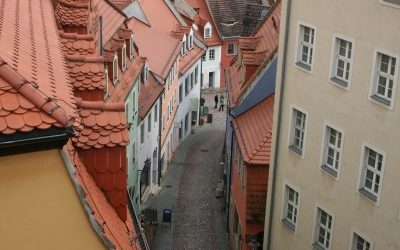 Blick von oben auf eine historische Altstadtgasse mit Kopfsteinpflaster, gesäumt von mehrstöckigen Häusern mit roten Ziegeldächern in traditioneller Bauweise.