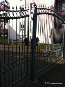 Eisernes Gartentor mit verzierungen und Spitzelementen vor einem rosa Gebäude mit Balkonen. Der Boden ist gepflastert. Das Tor ist leicht geöffnet und wirft Schatten auf den Boden. Im Hintergrund sind Fenster und ein Maschendrahtzaun zu sehen.