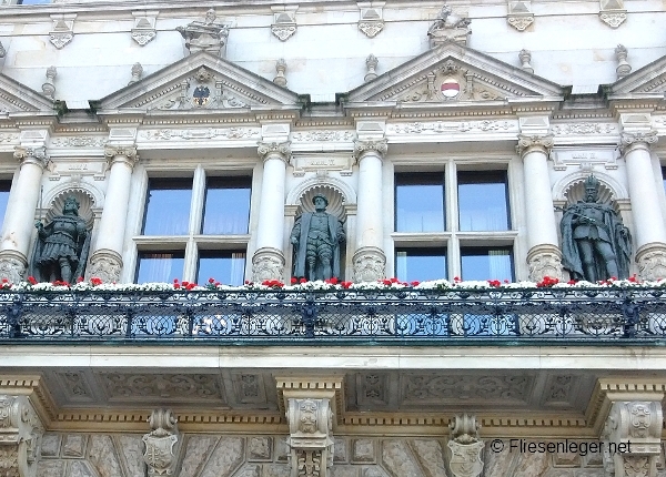 Historische Fassade mit mehreren Skulpturen zwischen Säulen. Darunter florale Verzierungen und ein kunstvoll gestaltetes Geländer. Die Architektur weist klassische Details und integrierte Fenster auf. Blumen schmücken den Balkon.