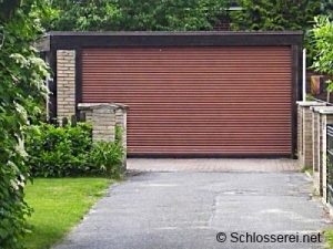 Eine Garage mit braunem Rolltor, umgeben von Zäunen und grüner Vegetation. Die Einfahrt ist asphaltiert. Schlichter rechteckiger Bau mit Ziegelwänden links.