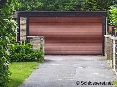Eine Garage mit braunem Rolltor, umgeben von Zäunen und grüner Vegetation. Die Einfahrt ist asphaltiert. Schlichter rechteckiger Bau mit Ziegelwänden links.