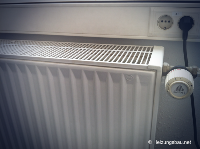 Ein weißer Heizkörper mit Thermostatventil in einem Innenraum. Neben dem Heizkörper befindet sich eine Steckdosenleiste mit zwei Steckdosen. Das Bild hat einen © Heizungsbau.net Vermerk.