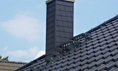 Modern gedecktes Satteldach mit dunklen glasierten Dachziegeln und einem rechteckigen, ebenfalls dunkel verkleideten Schornstein mit Metallabdeckung und Regenhaube. Im Vordergrund sind Schneefanggitter montiert, der Himmel ist leicht bewölkt.