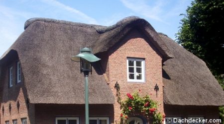 Backsteinhaus mit einem traditionell gedeckten Reetdach, das sich geschwungen an die Dachform anpasst. Vor dem Haus eine Straßenlaterne und eine blühende Kletterpflanze am Eingang.