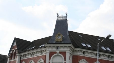 Historisches Gebäude mit aufwendig gestalteter Schieferdeckung auf einem steilen Turmdach. Die Fassade ist rot-weiß verputzt mit klassizistischen Fensterelementen, das Dach weist verschiedene Dachneigungen und kleine Gaubenfenster auf.