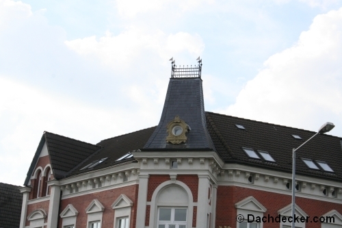Historisches Gebäude mit aufwendig gestalteter Schieferdeckung auf einem steilen Turmdach. Die Fassade ist rot-weiß verputzt mit klassizistischen Fensterelementen, das Dach weist verschiedene Dachneigungen und kleine Gaubenfenster auf.