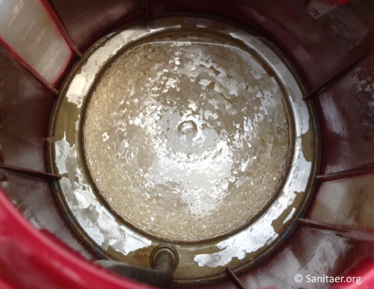Blick in das Innere eines Wasserboilers. Die Wände des Boilers sind mit Kalkablagerungen bedeckt, die eine raue, ungleichmäßige Oberfläche bilden. Der Rand des Boilers ist rostfarben, was auf eine metallische Struktur hinweist.