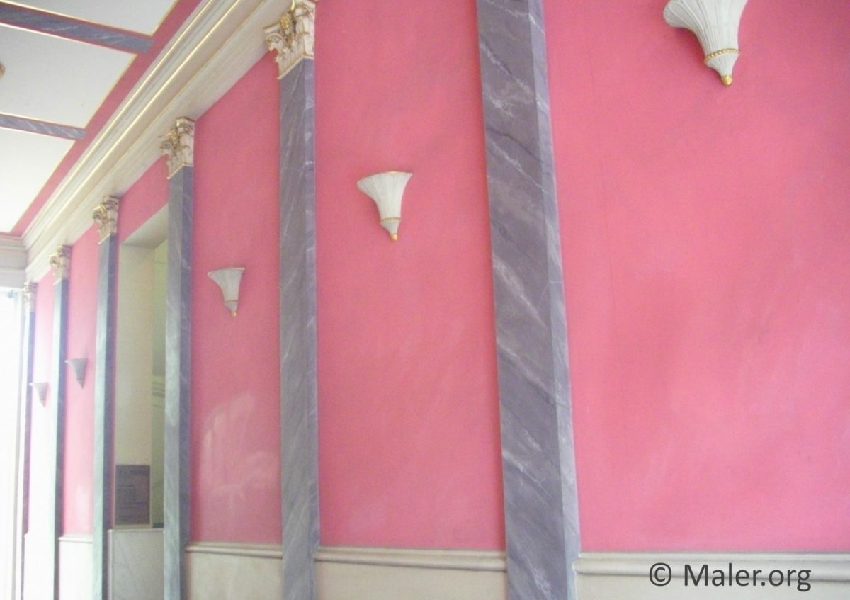Wand mit rosa Putz und grauen Marmorsäulen auf der rechten Seite. Es gibt drei weiße Wandlampen, die gleichmäßig verteilt sind. Die Decke hat goldene und blaue Details, während eine Lichtquelle von links kommt.