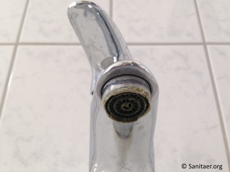 Nahaufnahme eines Wasserhahns mit Kalkablagerungen am Auslass. Der Wasserhahn ist aus silbernem Metall und vor einem weißen gefliesten Hintergrund angebracht.