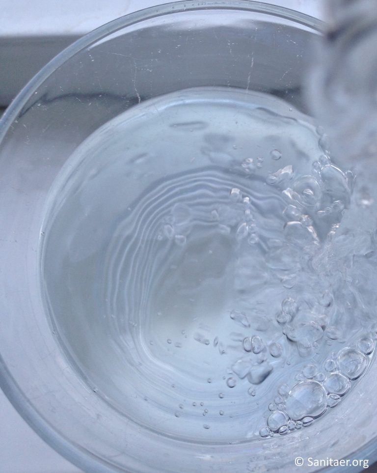 Ein Glas wird mit sprudelndem Wasser gefüllt. Das Wasser erzeugt Blasen und Wellen an der Oberfläche. Die Klarheit und Bewegung des Wassers sind deutlich sichtbar. Der Hintergrund ist unscharf.