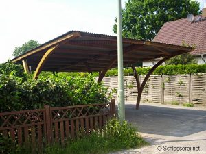 Ein offener Carport aus Holz mit gewölbter Dachkonstruktion steht neben einer Hecke. Im Vordergrund befindet sich ein Holzzaun, während im Hintergrund ein Haus mit Satteldach zu sehen ist. Ein Laternenpfahl steht an der Ecke des Carports.