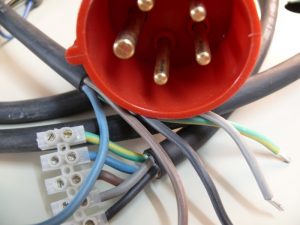Nahaufnahme eines roten CEE-Steckers mit sichtbaren Metallstiften. Mehrere bunte Stromkabel sind am Stecker befestigt und verlaufen durch eine weiße Lüsterklemme. Die Kabelisolierung zeigt verschiedene Farben, darunter blau, gelb-grün und grau.