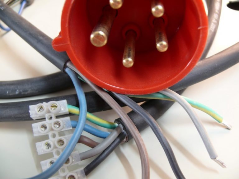 Nahaufnahme eines roten CEE-Steckers mit sichtbaren Metallstiften. Mehrere bunte Stromkabel sind am Stecker befestigt und verlaufen durch eine weiße Lüsterklemme. Die Kabelisolierung zeigt verschiedene Farben, darunter blau, gelb-grün und grau.