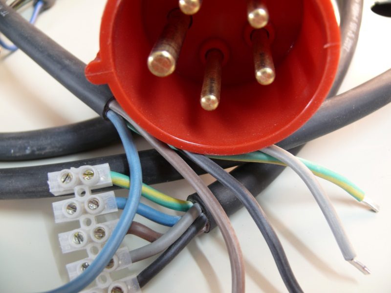 Nahaufnahme eines roten CEE-Steckers mit sichtbaren Metallstiften. Mehrere bunte Stromkabel sind am Stecker befestigt und verlaufen durch eine weiße Lüsterklemme. Die Kabelisolierung zeigt verschiedene Farben, darunter blau, gelb-grün und grau.