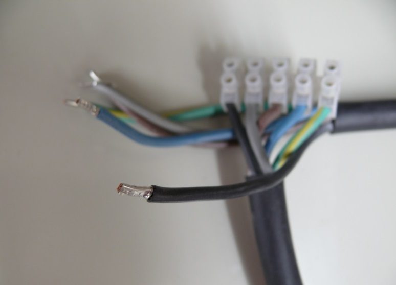 Nahaufnahme von mehreren abisolierten, farbigen Kabeln, die in einer weißen Lüsterklemme angeschlossen sind. Im Vordergrund ist ein schwarzes Kabel mit abisoliertem Ende sichtbar. Der Hintergrund ist unscharf, was den Fokus auf die Kabel legt.