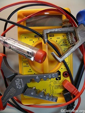 Ein gelbes Multimeter liegt zwischen verschiedenen Werkzeugen und Kabeln. Sichtbar sind ein Spannungsprüfer, Klemmen und ein Abisolierwerkzeug. Das Multimeter zeigt verschiedene Messbereiche an.