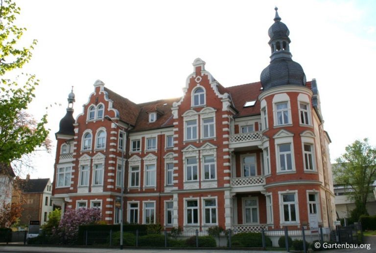 Ein historisches Gebäude mit roten Backsteinen und weißen Akzenten, verziert mit Türmchen und dekorativen Balkonen. Es hat mehrere große Fenster und ein rotes Dach. Im Vordergrund sind blühende Büsche zu sehen.