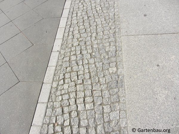 Kopfsteinpflaster aus Granit, eingefasst von glatten Plattenbelägen – Übergang zwischen zwei unterschiedlichen Gehwegoberflächen im urbanen Raum.