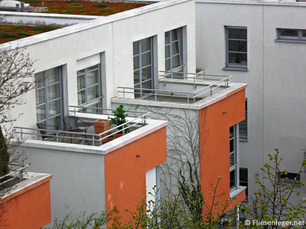 Moderne Wohnanlage mit orange-weißen Fassaden und kleinen Balkonen. Zwei Balkone sind mit Pflanzen und Möbeln dekoriert. Es gibt mehrere große Fenster, die zu den Wohnungen gehören. Einige Bäume und Sträucher sind im Vordergrund sichtbar.