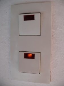 Zwei weiße Wandschalter auf einer rechteckigen Platte. Der obere Schalter ist aus, während der untere Schalter eingeschaltet ist und ein kleines rotes Licht leuchtet. Die Hintergrundwand ist weiß und leicht strukturiert.