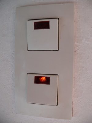 Zwei weiße Wandschalter auf einer rechteckigen Platte. Der obere Schalter ist aus, während der untere Schalter eingeschaltet ist und ein kleines rotes Licht leuchtet. Die Hintergrundwand ist weiß und leicht strukturiert.