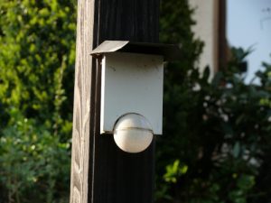 Eine rechteckige Außenleuchte mit weißem Gehäuse und einer runden Glaskuppel ist an einem dunklen Holzpfosten montiert. Im Hintergrund sind grüne Büsche und ein Teil eines Gebäudes zu sehen. Natürliche Beleuchtung zeigt Detailreichtum.
