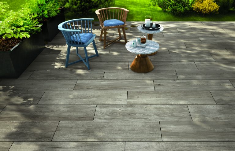 Großformatige Terrassenplatten in Holzoptik auf einer modernen Gartenterrasse mit zwei runden Tischen, zwei Stühlen und üppiger grüner Bepflanzung im Hintergrund.