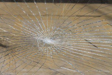 Zersprungenes Glas mit einem zentralen Einschlagpunkt, von dem aus zahlreiche Risse radial nach außen verlaufen. Der Hintergrund ist verschwommen und beige.