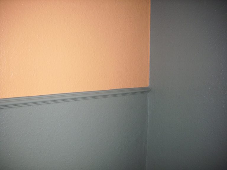 Ecke eines Raumes mit zweifarbiger Wand: oben in hellem Orange und unten in dunklem Grau. Ein schmaler, grauer Zierstreifen trennt die beiden Farbbereiche. Die Textur der Wand ist glatt.