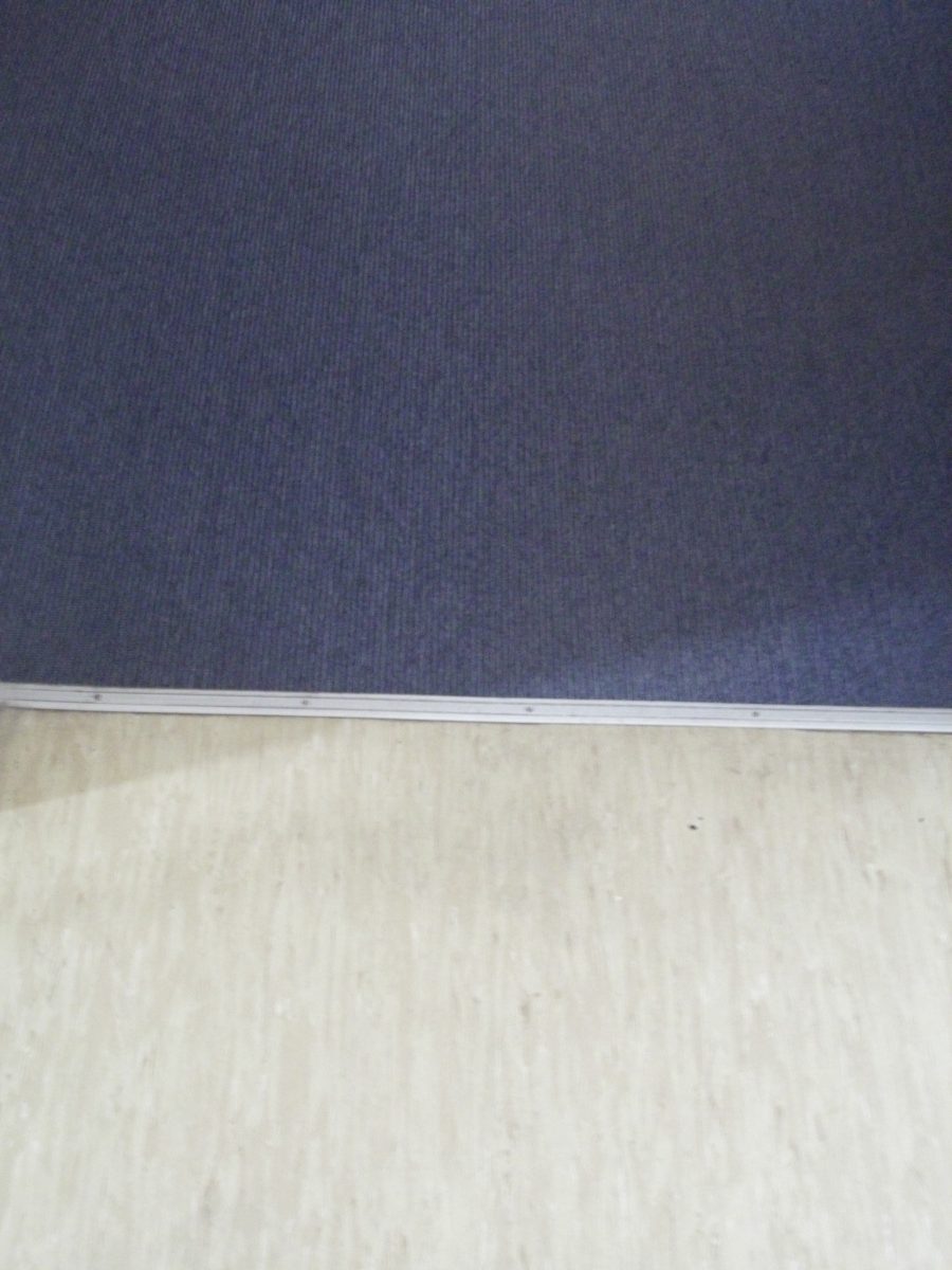 Dunkler Teppich grenzt an einen hellen, glatten Bodenbelag. Die beiden Materialien sind durch eine schmale Metallleiste verbunden. Der Teppich ist blau, während der Boden beige ist. Diese Kombination könnte in einem Büro oder Flur zu finden sein.