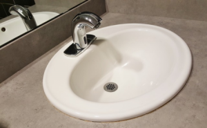 Ein weißes Waschbecken mit einem modernen, silbernen Wasserhahn, eingebaut in eine graue Arbeitsplatte. Im Hintergrund ist ein Spiegel zu sehen, der das Waschbecken und die Umgebung reflektiert.