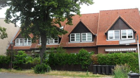 Reihenhaus mit rotem Satteldach, weißen Fenstern und Vorgarten, von einem großen Baum und Hecken umgeben.