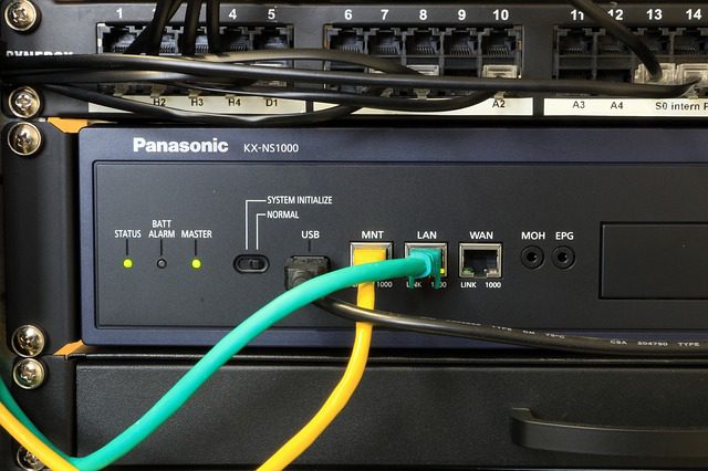 Nahaufnahme einer Panasonic KX-NS1000 Kommunikationsanlage in einem Serverrack. Verschiedene Kabel sind angeschlossen, darunter ein gelbes und ein grünes. LED-Anzeigen leuchten, darunter "Status" und "BATT". Oben ein Patchpanel mit Ports.