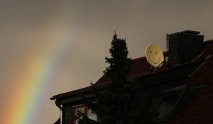 Dach eines Wohnhauses mit einer gelben Satellitenschüssel, auf der ein Smiley-Gesicht zu sehen ist. Im Hintergrund ist ein Regenbogen am bewölkten Himmel sichtbar.