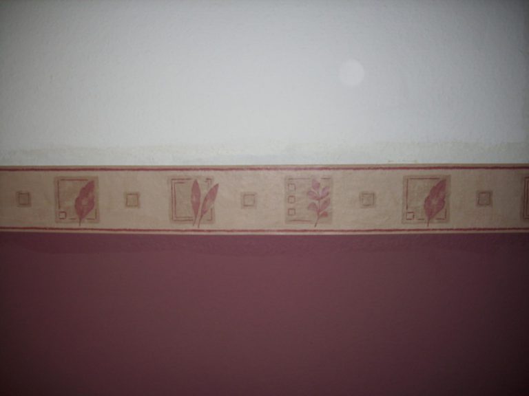 Tapete mit Floralmuster: Die Wand zeigt eine Tapete mit Bordüre, auf der abwechselnd Blätter und geometrische Formen abgebildet sind. Die Tapete hat einen zweifarbigen Hintergrund, oben in Weiß und unten in Beige.
