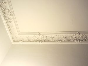 Eine weiße Zimmerdecke mit detaillierten Stuckverzierungen entlang der Kante. Die Ornamente sind symmetrisch und kunstvoll gestaltet, verleihen dem Raum einen eleganten, klassischen Look.
