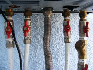 Verschiedene Rohre und Ventile sind an einer Wand befestigt. Es gibt fünf weiße und ein flexibles Metallrohr. Vier rote Ventile dienen zur Steuerung des Wasserflusses. Die Wand ist hellblau gestrichen.