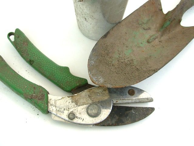 Gartenwerkzeug-Set mit einer schmutzigen Handschaufel, einer Schere mit grünem Griff und einem Metallblumentopf auf weißem Hintergrund. Die Werkzeuge zeigen Gebrauchsspuren und sind staubig und rostig.