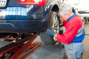 Kfz-Mechaniker in roter Arbeitskleidung prüft ein Hinterrad an einem blauen Fahrzeug auf einer Hebebühne in einer Werkstatt, mit Fokus auf Reifen und Fahrwerksbereich.
