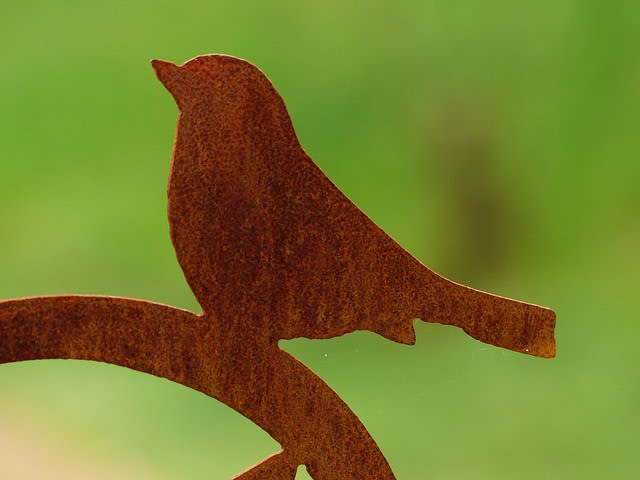 Scherenschnitt einer Vogelsilhouette aus rostigem Metall vor einem verschwommenen grünen Hintergrund. Der Vogel sitzt auf einem leicht gebogenen Zweig, die Details sind einfach gehalten, was einen minimalistischen Eindruck erzeugt.