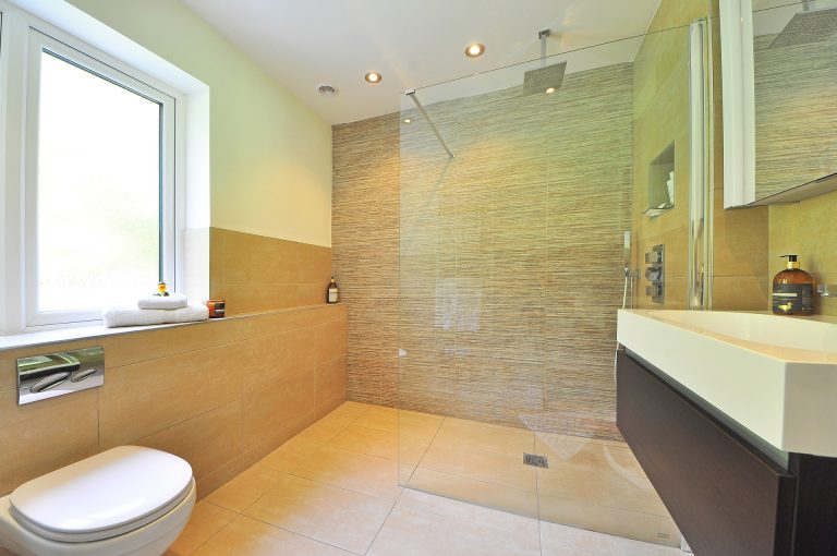 Modernes Badezimmer mit beiger Fliesenwand und großer, ebenerdiger Dusche mit Glasabtrennung. Links ist ein Fenster zu sehen, davor ein WC. Auf dem Waschbecken stehen Pflegeprodukte. Deckenleuchten sorgen für Beleuchtung.