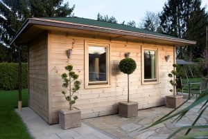 Holzhaus mit zwei Fenstern, flankiert von zwei symmetrischen Pflanzen in quadratischen Töpfen. Das Gebäude steht auf einer gepflasterten Fläche, umgeben von Rasen und Bäumen im Hintergrund. Rustikaler Stil mit grüner Dachabdeckung.