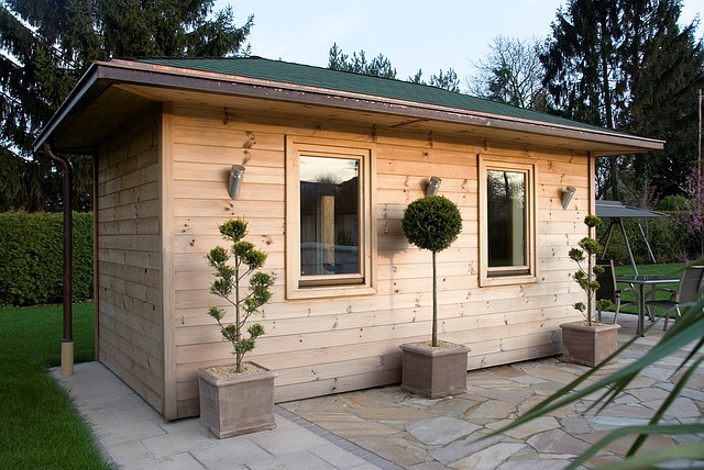 Holzhaus mit zwei Fenstern, flankiert von zwei symmetrischen Pflanzen in quadratischen Töpfen. Das Gebäude steht auf einer gepflasterten Fläche, umgeben von Rasen und Bäumen im Hintergrund. Rustikaler Stil mit grüner Dachabdeckung.