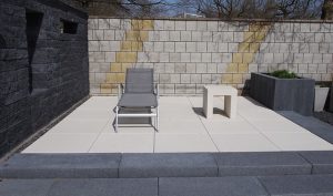 Ein grauer Liegestuhl steht auf einer gepflasterten Terrasse neben einem kleinen, weißen Beistelltisch. Dahinter eine hohe Mauer aus gemusterten Betonziegeln. Rechts davon sind in einem grauen Pflanzkübel grüne Pflanzen zu sehen.