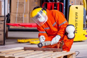 Eine Person in Schutzkleidung und Helm arbeitet mit einem Winkelschleifer in einer Werkstatt. Funken fliegen von dem Metallstück, das sie bearbeiten. Im Hintergrund sind Kartons und ein gelber Gabelstapler zu sehen.