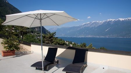 Flachdachterrasse mit zwei modernen Sonnenliegen und einem weißen Sonnenschirm, umgeben von mediterraner Landschaft mit Blick auf einen See und schneebedeckte Berge im Hintergrund.