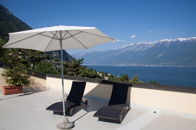 Flachdachterrasse mit zwei modernen Sonnenliegen und einem weißen Sonnenschirm, umgeben von mediterraner Landschaft mit Blick auf einen See und schneebedeckte Berge im Hintergrund.